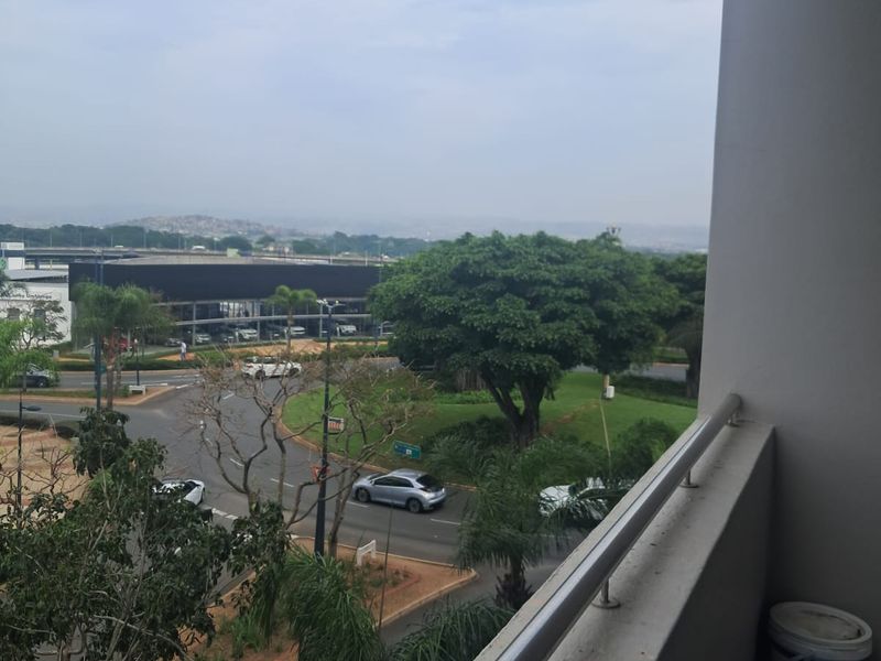 An elevated view from a balcony overlooking a suburban street intersection, landscaped greenery, and surrounding commercial buildings.