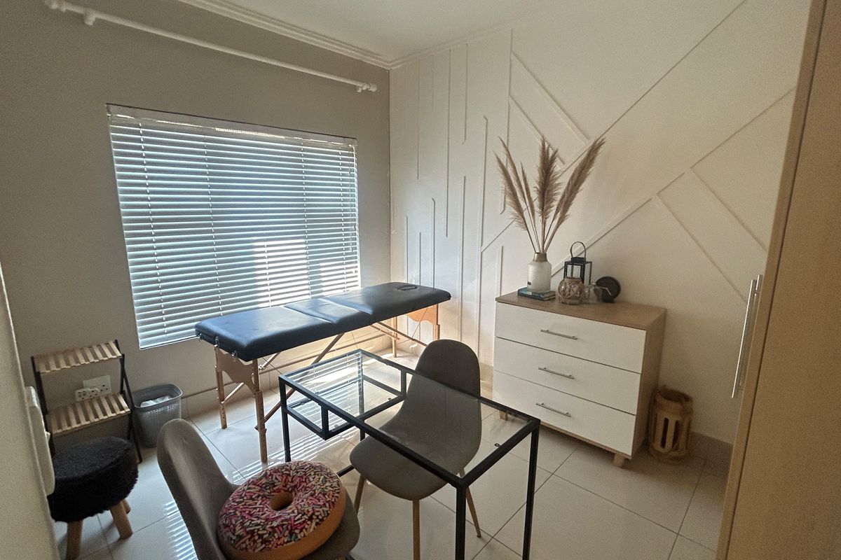 A well-lit bedroom featuring modern wall paneling, neutral tiled flooring, and venetian blinds, currently utilized as a functional workspace or treatment room.