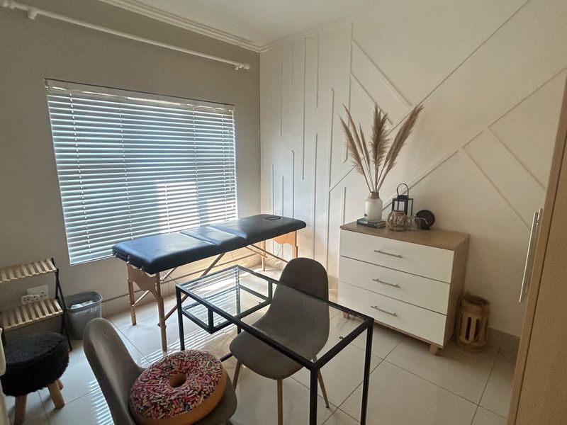 A well-lit bedroom featuring modern wall paneling, neutral tiled flooring, and venetian blinds, currently utilized as a functional workspace or treatment room.