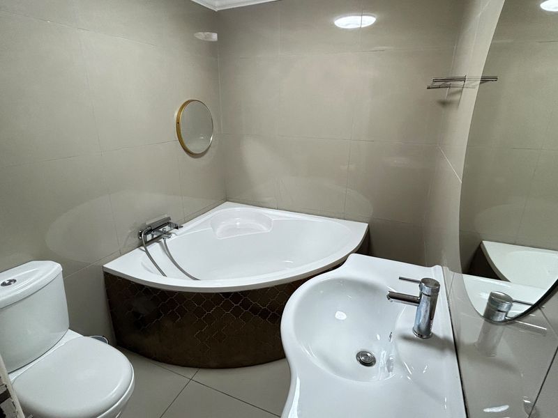 A clean and modern bathroom featuring a corner bathtub, pedestal washbasin, toilet, and neutral wall tiling.