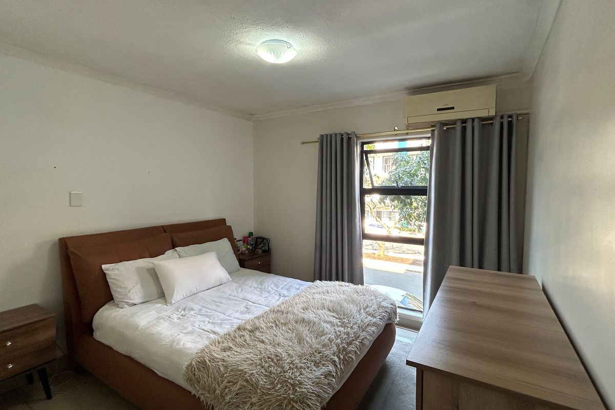 A well-lit bedroom featuring a wall-mounted air conditioning unit and a large window with curtains, providing natural light and ventilation.