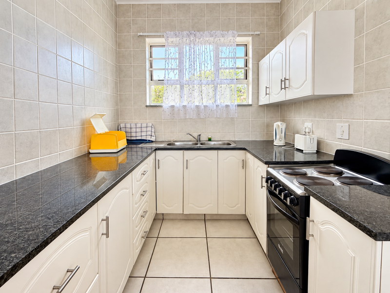 A functional kitchen featuring granite countertops, white cabinetry, a stainless steel double sink, and a freestanding electric stove with tiled walls and flooring.
