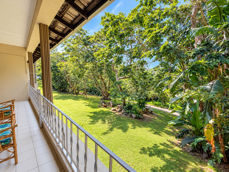 A spacious covered balcony featuring tiled flooring and a metal balustrade, offering a pleasant view over the lush green garden and mature trees.