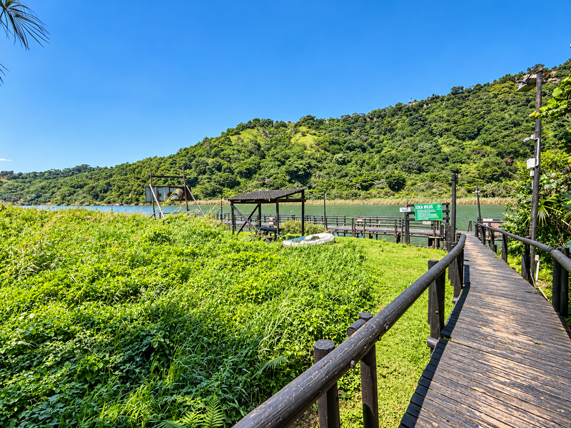 Scenic view of a wooden boardwalk leading to a jetty and covered pier structure overlooking a river and lush green hillside.