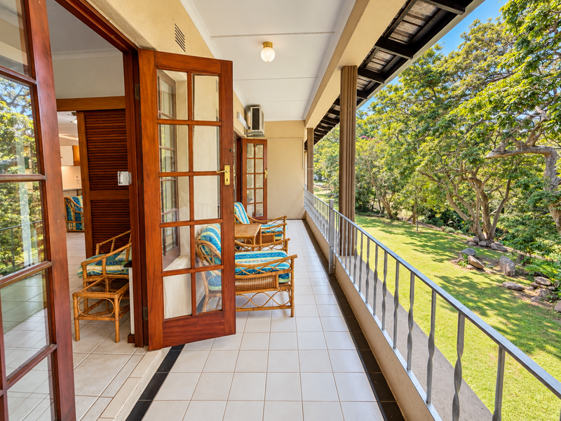 A spacious covered balcony featuring tiled flooring, a metal balustrade, and wooden framed glass doors, overlooking a lush green garden.