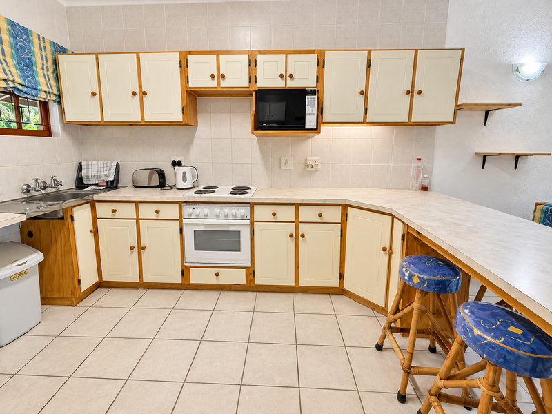 A functional kitchen featuring dated timber-trimmed cabinetry, laminate countertops, a built-in oven and hob, and neutral tiled flooring with a breakfast bar area.