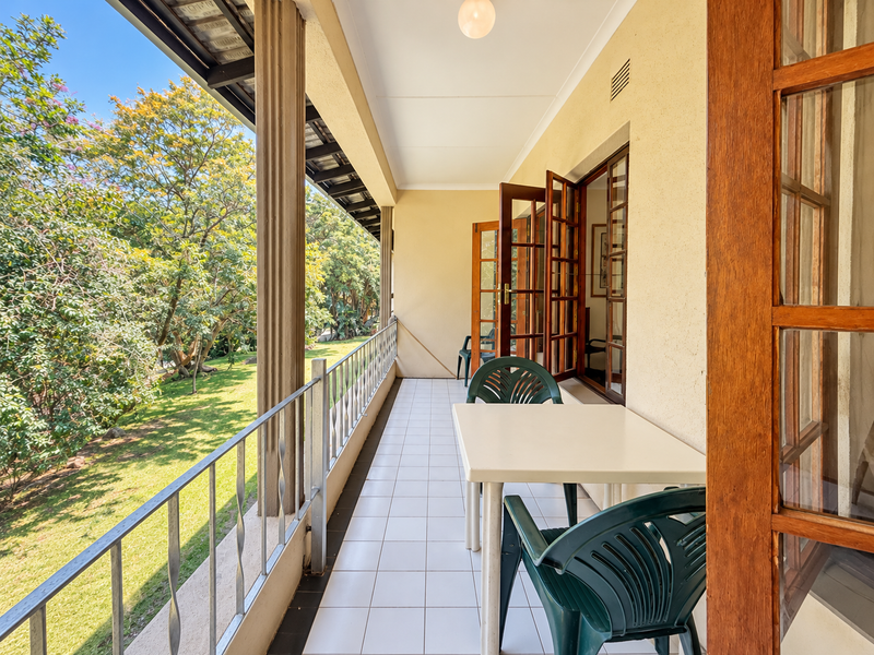 A covered balcony featuring tiled flooring, metal balustrades, and wooden framed glass doors, offering a pleasant view of the surrounding garden area.