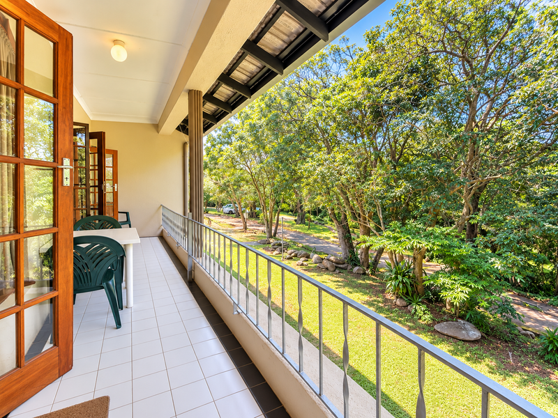 A spacious covered balcony featuring tiled flooring and a metal balustrade, offering a pleasant view of the surrounding trees and garden area.