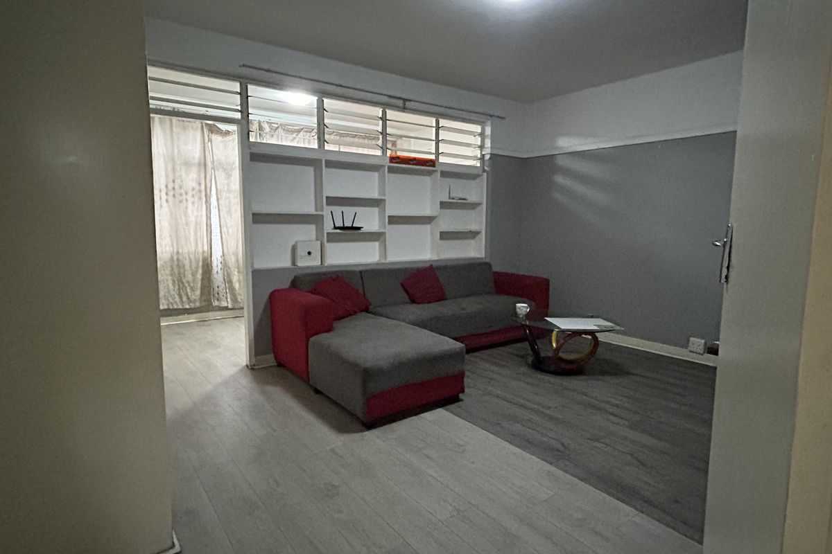 A living room featuring laminate flooring, a built-in shelving unit, and louvre windows, offering a functional space with potential for modern updates.
