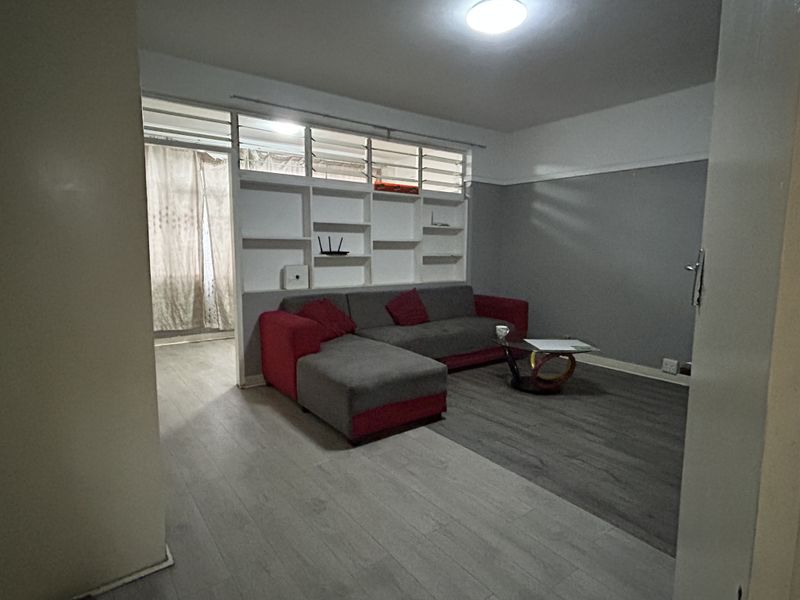 A living room featuring laminate flooring, a built-in shelving unit, and louvre windows, offering a functional space with potential for modern updates.