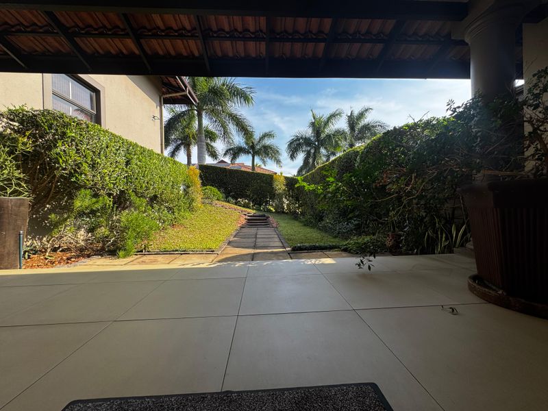 A covered patio area featuring tiled flooring and concrete columns, overlooking a well-maintained garden with a stone pathway and established hedges.