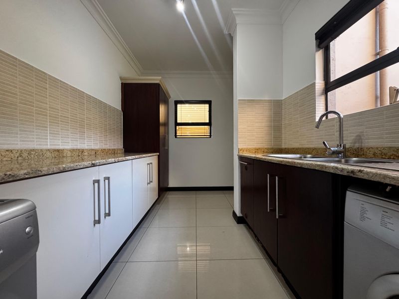 A clean and functional scullery featuring granite countertops, dark and white cabinetry, a stainless steel sink, and tiled finishes.