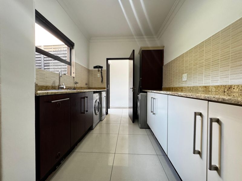 A functional scullery featuring granite countertops, ample built-in cabinetry, a stainless steel sink, and tiled finishes, providing efficient space for laundry and kitchen prep.