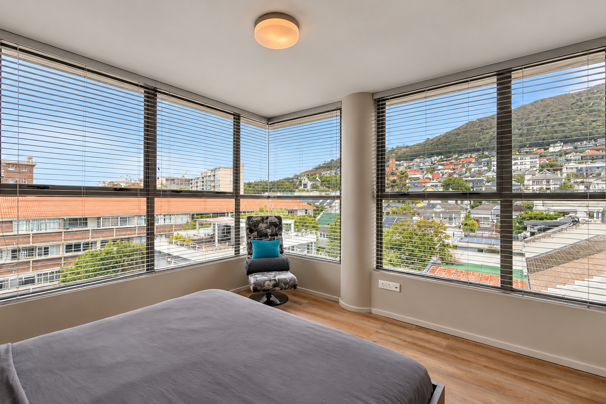 A bright bedroom featuring large corner windows with venetian blinds, offering expansive urban and mountain views, complemented by modern laminate flooring.
