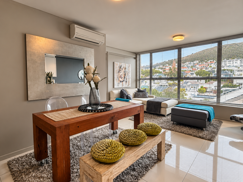 A bright open-plan living and dining area featuring large windows with expansive city and mountain views, tiled floors, and a wall-mounted air conditioning unit.