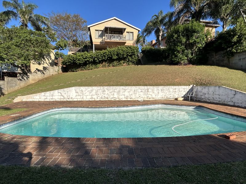 A backyard pool area featuring a swimming pool with brick paving, a retaining wall, and a view of the elevated house with a balcony.