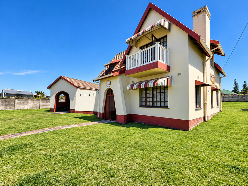 A well-maintained two-story house featuring a distinctive architectural style, a balcony, striped awnings, a chimney, and an attached carport set on a grassy lot.