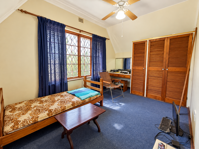 A bedroom featuring a built-in wooden wardrobe, vanity, ceiling fan, and leaded glass windows, offering a functional space with potential for modernization.