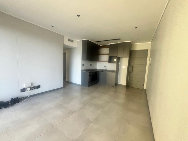 An open-plan living and kitchen area featuring modern dark cabinetry, integrated oven, tiled flooring, and recessed lighting in a clean, neutral space.