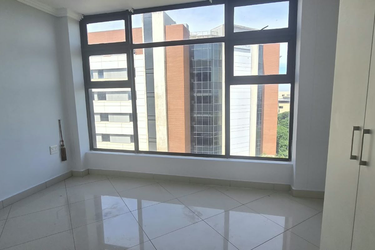 A bright, empty bedroom featuring large aluminium-framed windows with urban views, tiled flooring, and a built-in wardrobe.