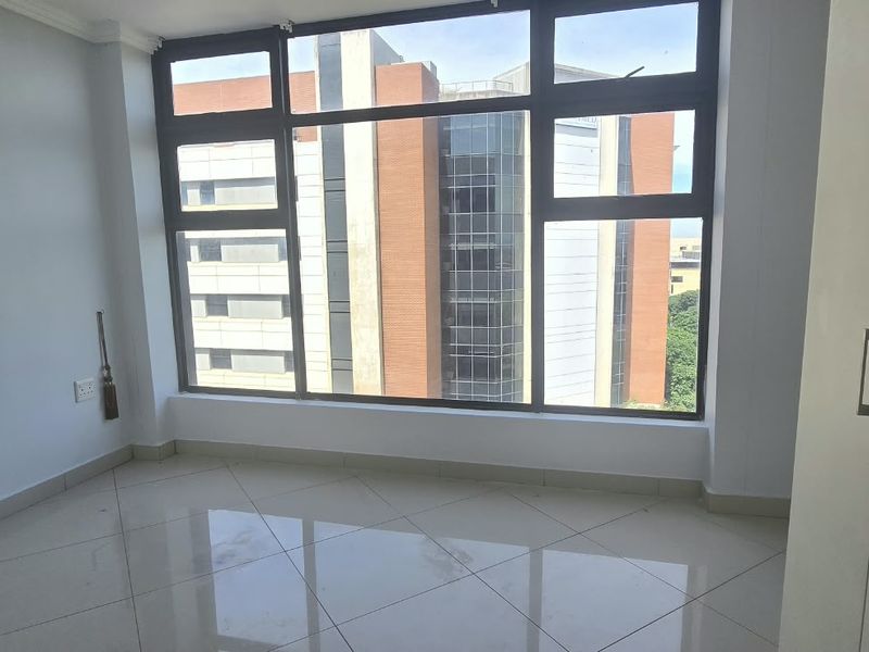 A bright, empty bedroom featuring large aluminium-framed windows with urban views, tiled flooring, and a built-in wardrobe.