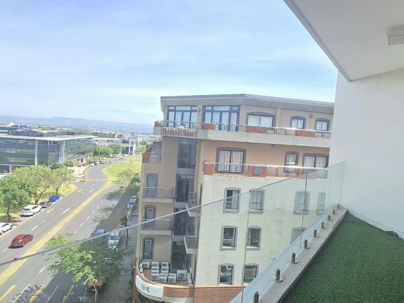 A modern balcony featuring a glass balustrade, artificial grass flooring, and expansive views of the surrounding urban landscape.