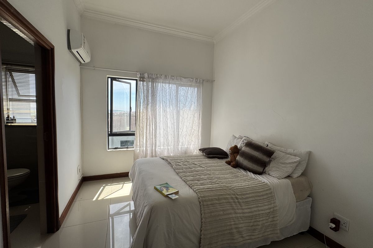A bright bedroom featuring tiled flooring, cornice detailing, and a wall-mounted air conditioning unit, with an adjoining bathroom visible through the doorway.