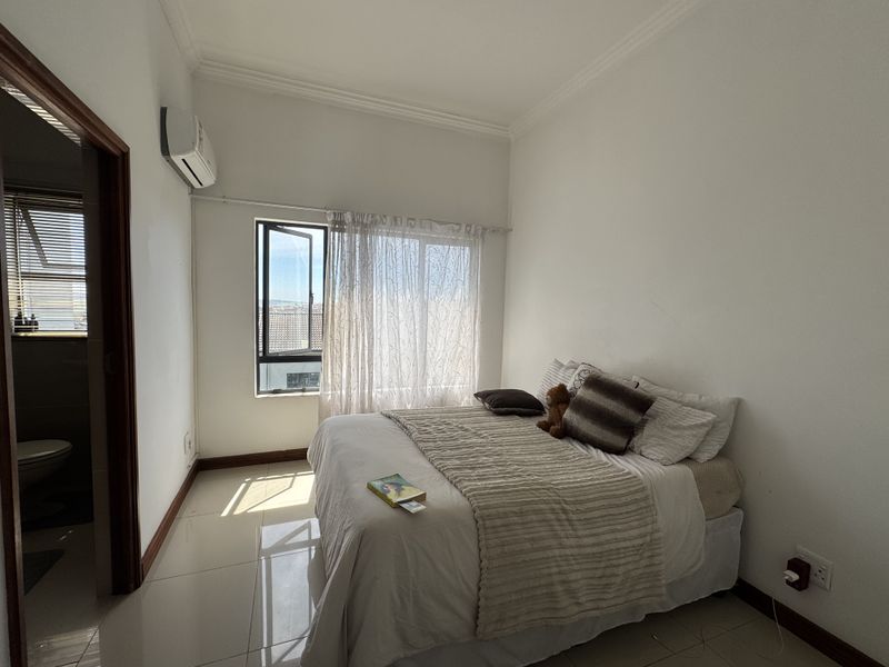 A bright bedroom featuring tiled flooring, cornice detailing, and a wall-mounted air conditioning unit, with an adjoining bathroom visible through the doorway.