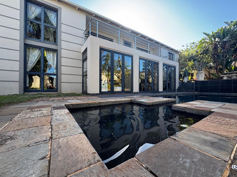 Rear exterior view featuring a swimming pool with stone surround, leading to a modern home with glass folding doors and an upper-level balcony.