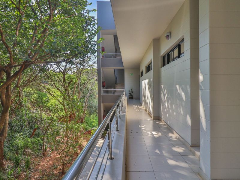 An open-air apartment building walkway featuring tiled flooring, a modern stainless steel balustrade, and views of the surrounding greenery.