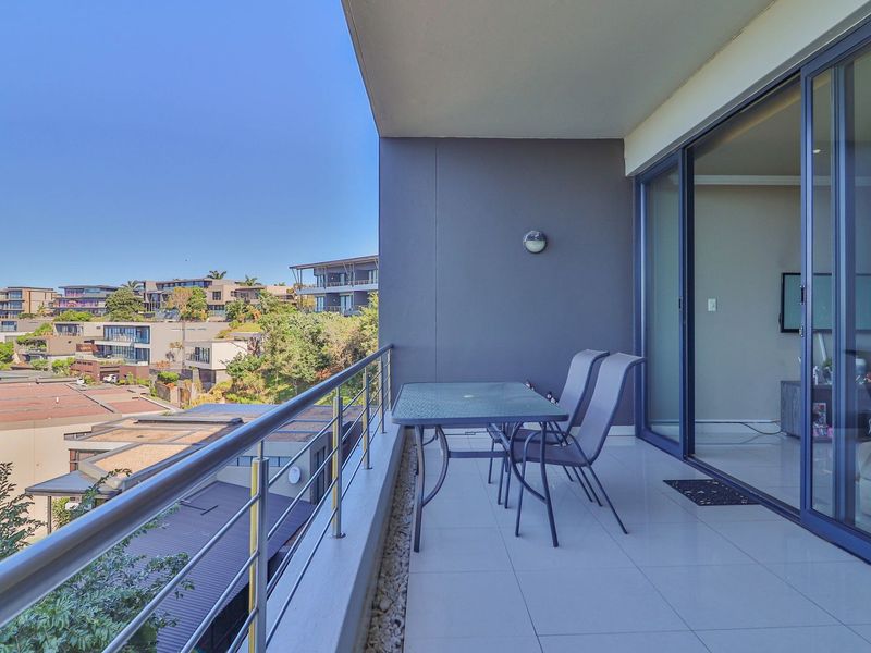 A spacious covered balcony featuring tiled flooring, a modern stainless steel balustrade, and large sliding glass doors leading into the apartment.