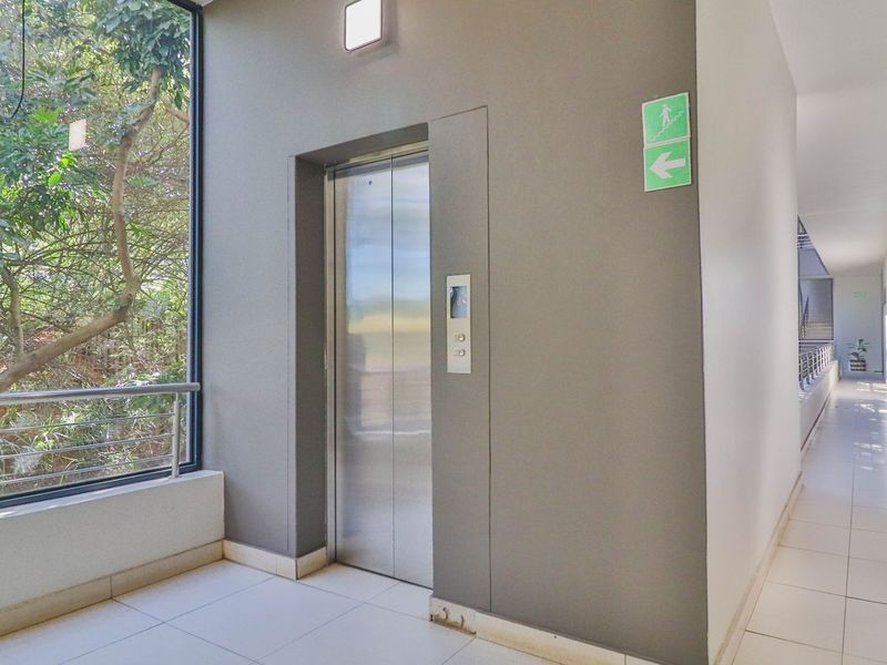A clean, modern apartment building hallway featuring tiled flooring, a stainless steel elevator, and a large window overlooking greenery.