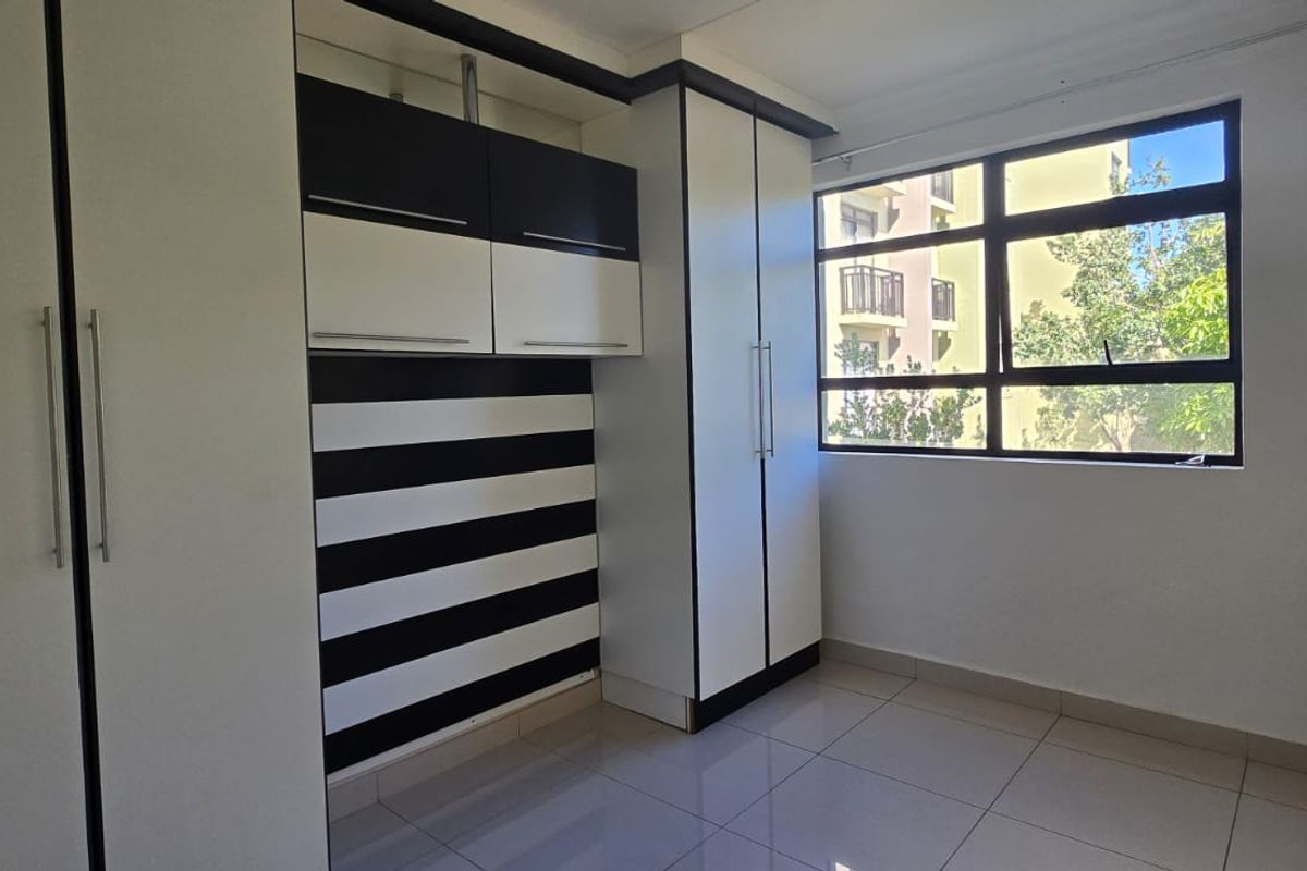 A bright, empty bedroom featuring modern built-in cupboards with a black and white finish, tiled flooring, and a large window providing natural light.