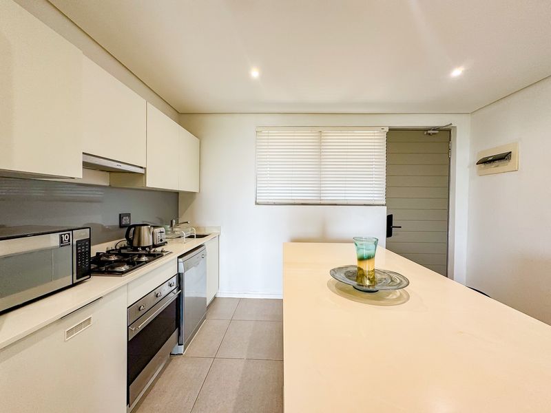 Modern kitchen featuring sleek white cabinetry, stone countertops, a gas hob, integrated appliances, and a central island, finished with neutral floor tiles.