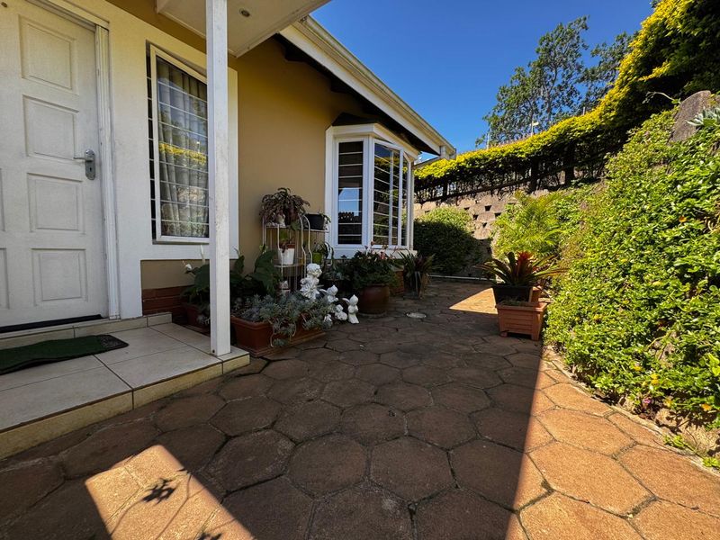 A paved outdoor patio area featuring a bay window, security bars, and a stone retaining wall in a landscaped garden setting.