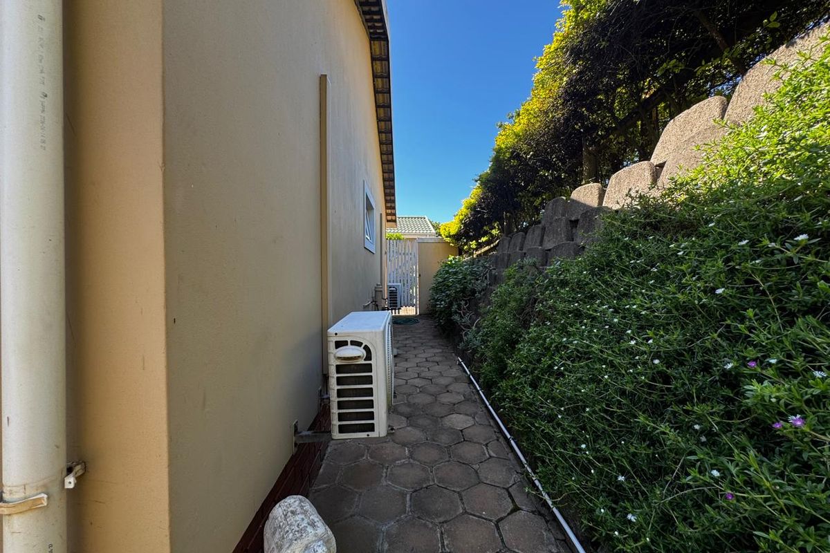 A side exterior walkway featuring paved ground, a retaining wall with landscaping, and an external air conditioning unit.
