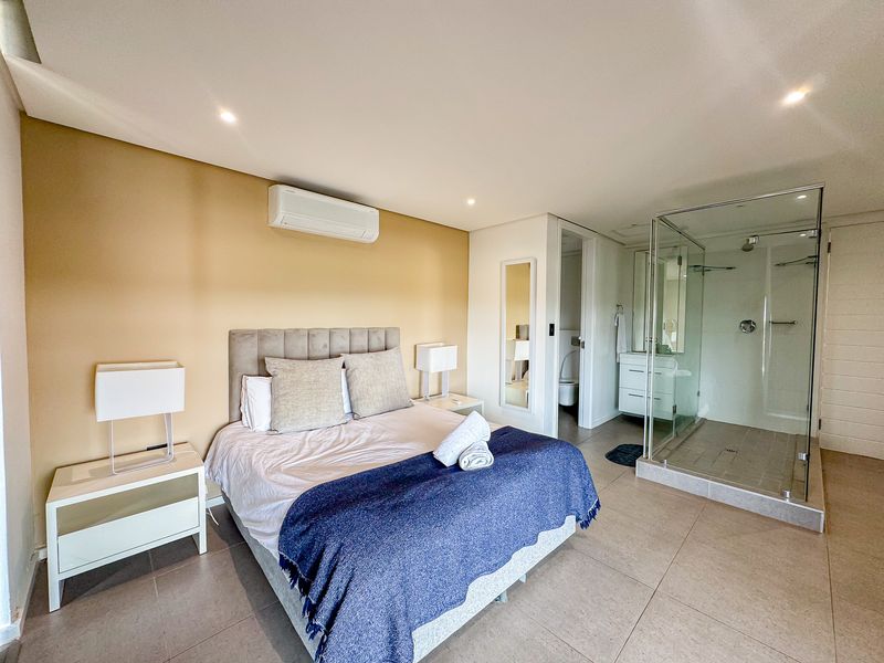 A modern bedroom featuring tiled flooring, an integrated air conditioning unit, and an en-suite bathroom with a glass-enclosed shower and vanity.