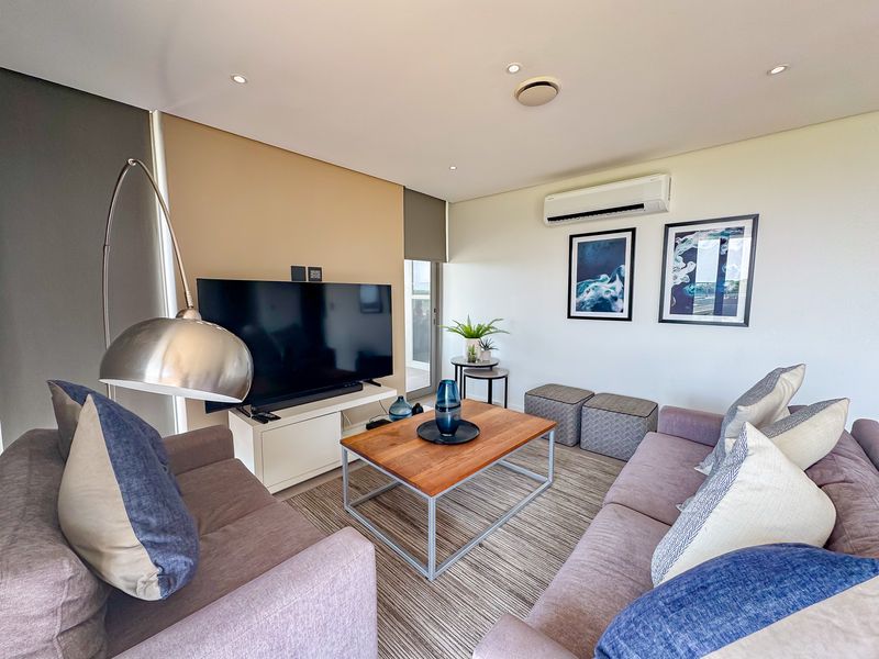 A well-maintained living room featuring recessed downlighting, a wall-mounted split-system air conditioner, and neutral wall finishes with roller blinds.