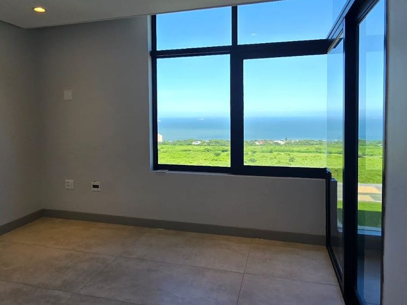A bright, empty bedroom featuring large format floor tiles, recessed lighting, and expansive windows offering a scenic coastal view.