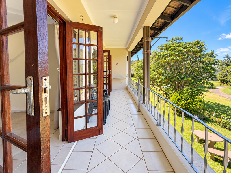A spacious covered balcony featuring tiled flooring, metal railings, and wooden french doors, offering a pleasant view of the surrounding garden area.