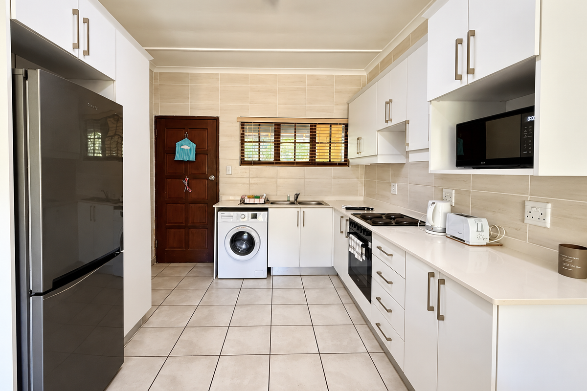 A clean and functional kitchen featuring white cabinetry, stone countertops, built-in oven, hob, and an integrated washing machine with neutral tiled finishes.