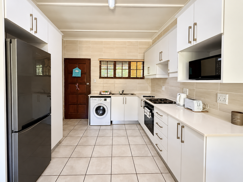 A clean and functional kitchen featuring white cabinetry, stone countertops, built-in oven, hob, and an integrated washing machine with neutral tiled finishes.