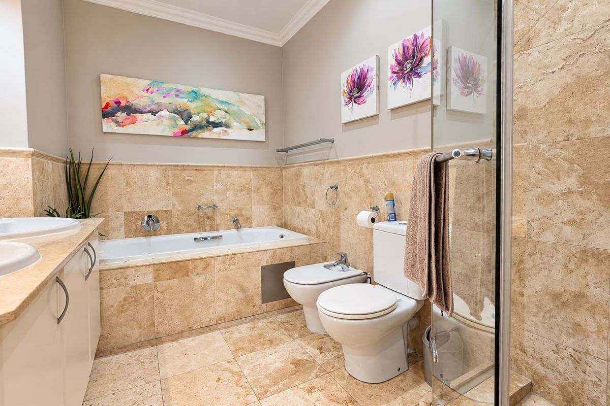 A well-appointed bathroom featuring a double vanity, built-in bathtub, bidet, toilet, and a glass-enclosed shower, finished with elegant travertine tiling throughout.
