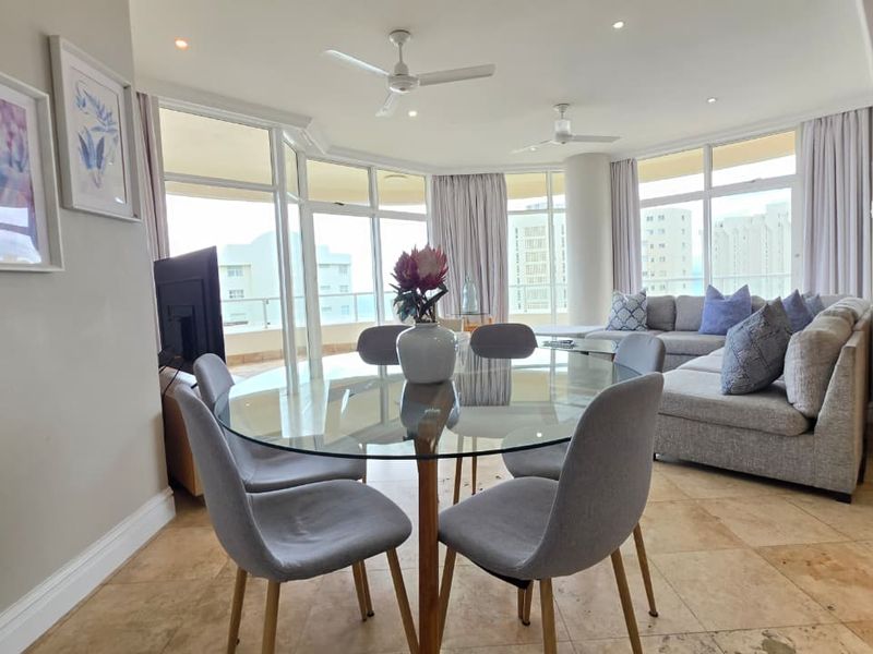 Spacious open-plan living and dining area featuring natural stone flooring, recessed lighting, ceiling fans, and expansive glass doors leading to a private balcony.