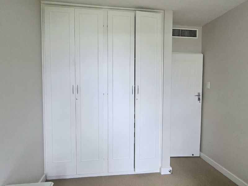 A clean, empty bedroom featuring white built-in cupboards, neutral wall finishes, and an integrated air conditioning vent.