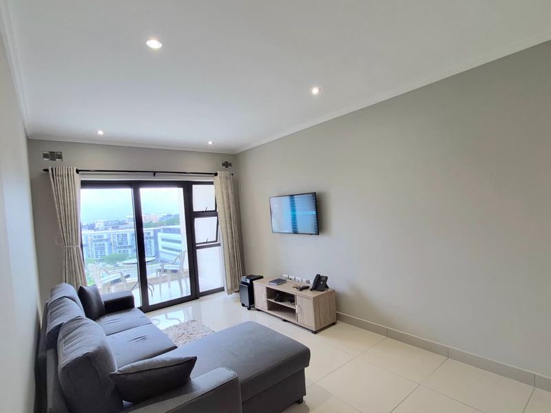 A bright and modern living room featuring neutral tiled flooring, recessed downlighting, and large sliding glass doors leading to an outdoor balcony area.
