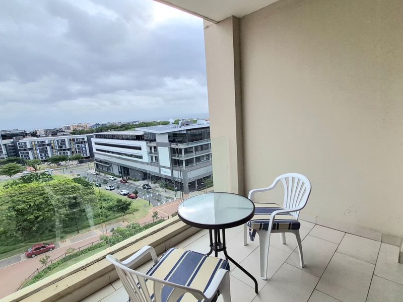 A covered balcony featuring a glass balustrade and tiled flooring, offering an elevated view of the surrounding urban landscape.