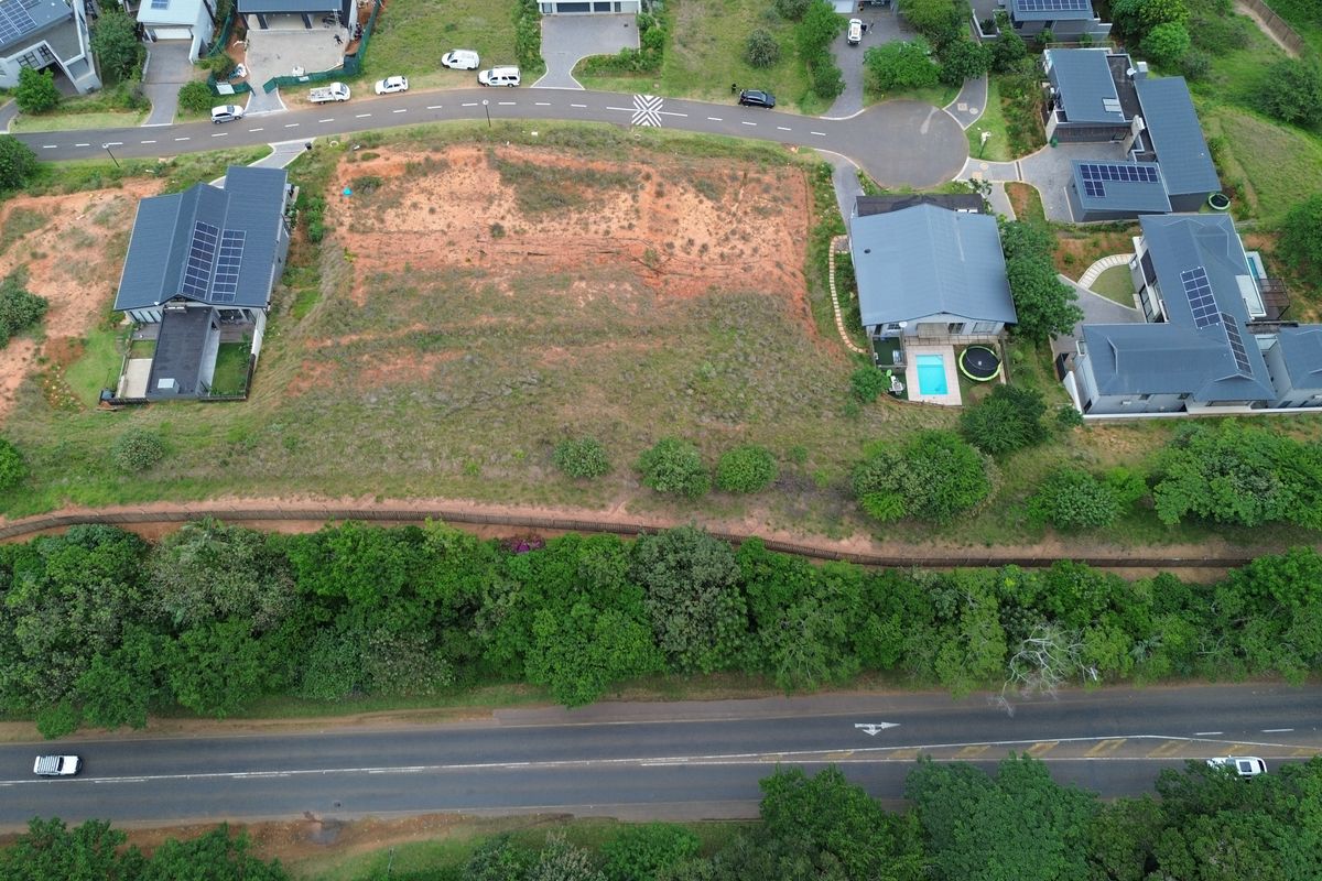 Aerial view of a vacant residential plot situated within a developed estate, featuring natural terrain and bordering a retaining wall.