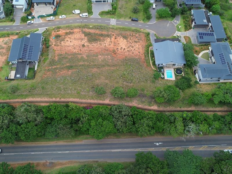 Aerial view of a vacant residential plot situated within a developed estate, featuring natural terrain and bordering a retaining wall.
