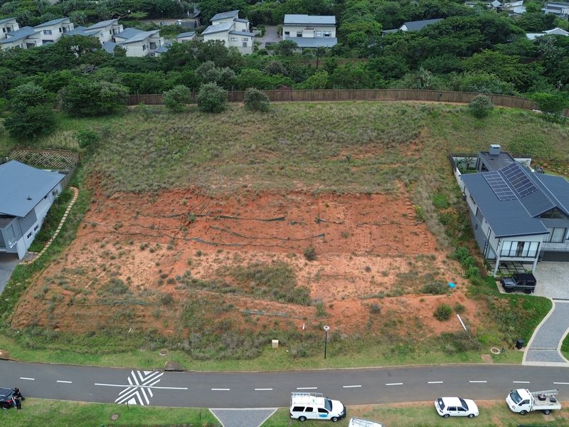 An aerial view of a vacant residential plot situated between existing modern homes, featuring natural vegetation and street access.